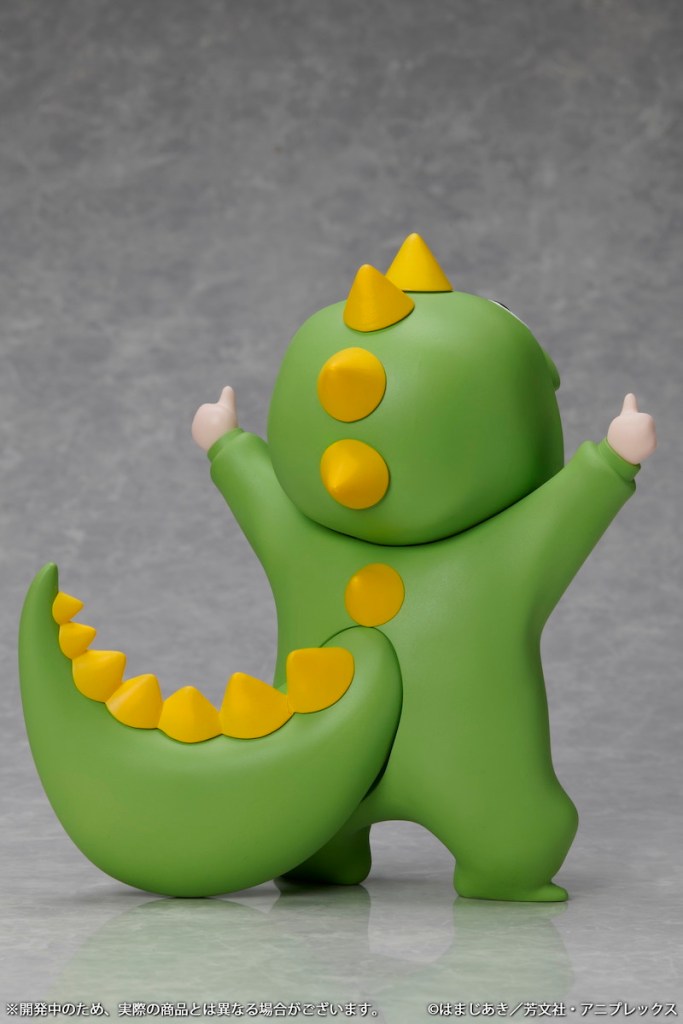 A cute green figure resembling a dinosaur, designed with spikes and a large tail, standing upright with both arms raised in excitement.
